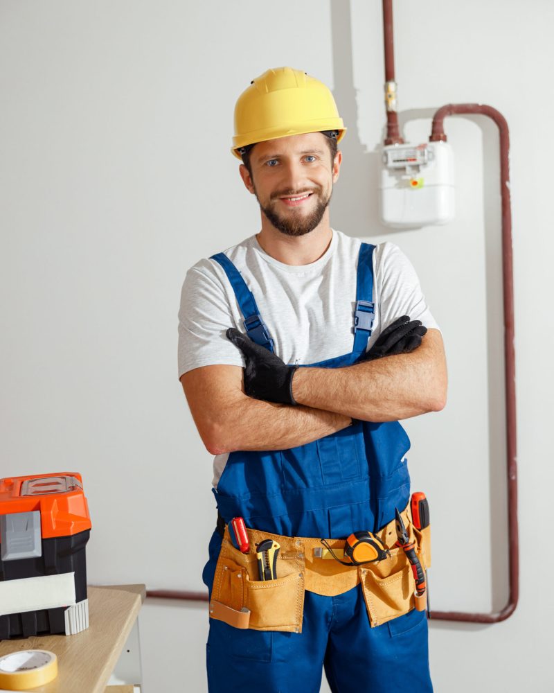 Professional electrician technician in uniform, hard hat and protective gloves smiling at camera while standing with arms crossed indoors. Manual work, maintenance, occupation concept